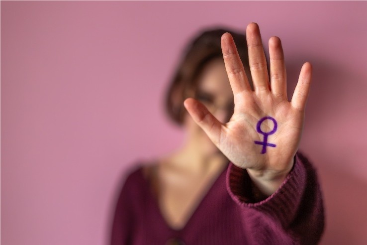 Una mujer sobre fondo borroso apunta a la cámara con la mano extendida, en cuya palma está dibujado el símbolo femenino de color morado, como muestra de rechazo a la violencia.